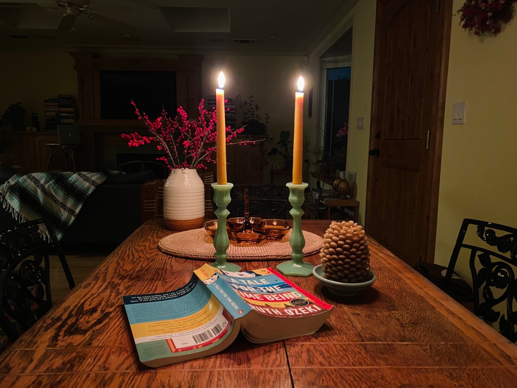Kitchen table — wood, worn, loved — holds two jade-green candlesticks with yellow hand-dipped beeswax candles, lit; a vase of red winter berry branches; a beeswax candle shaped like a pinecone; a woven grass placemat; glass amber-colored decorative dish for olives or pistachios; and a book, splayed open with the cover visible: A Tale for the Time Being by Ruth Ozeki.