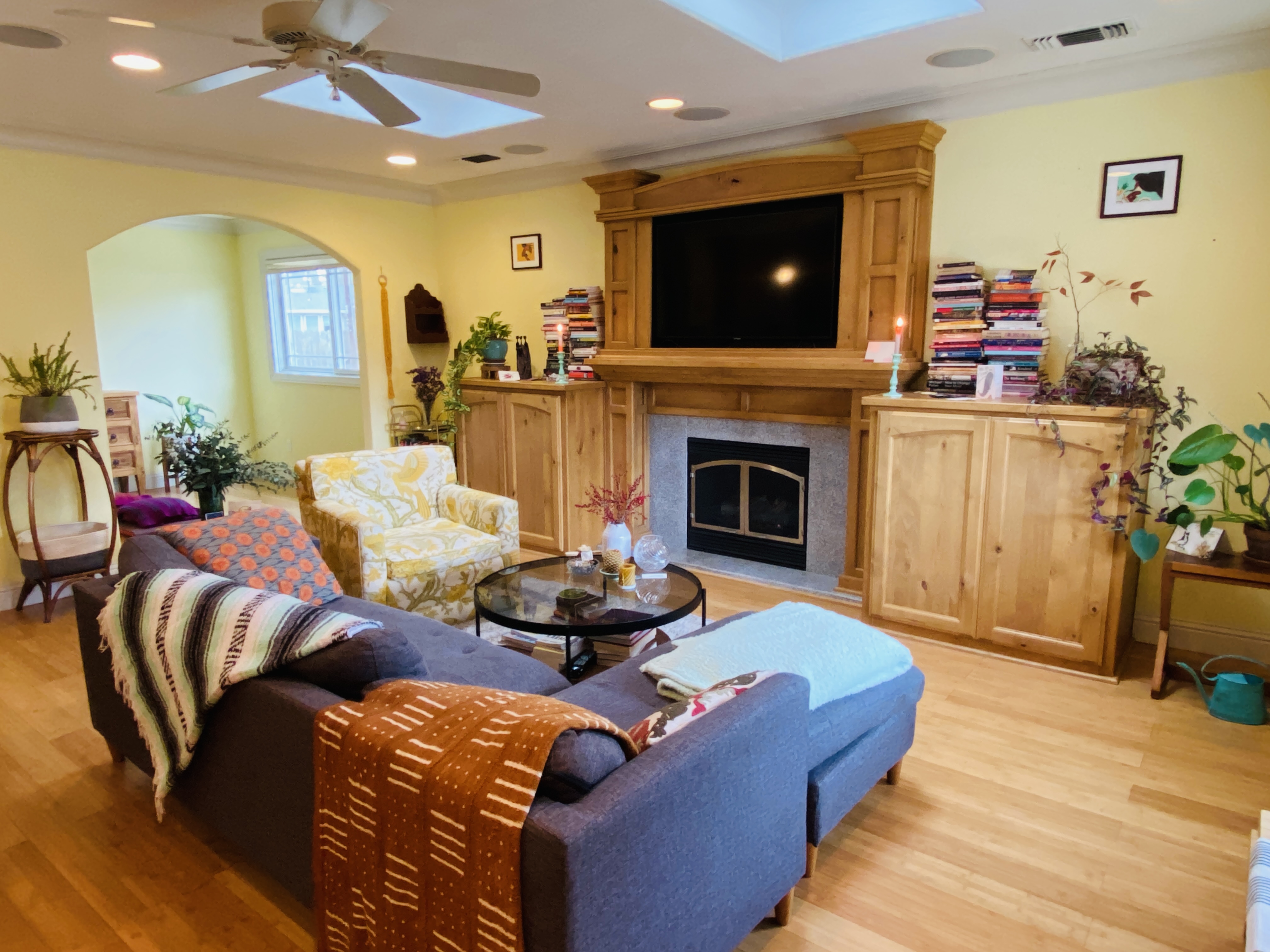 A photo of Katie's living room: soft yellow walls, lots of built-in wood, bamboo floors, a couch with many colorful blankets and throw pillows, a yellow patterned armchairs, some houseplants, a central big-screen tv, and stacks of books.
